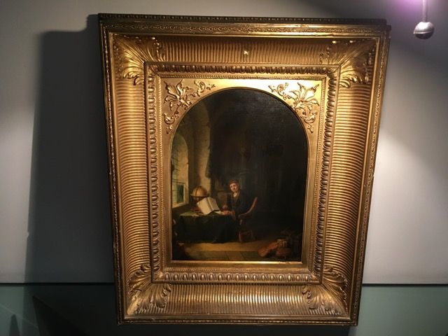 Une peinture à l'huile cintrée, encadrée d'un grand cadre doré orné, représentant un personnage lisant dans un bureau aux murs de pierre faiblement éclairé.