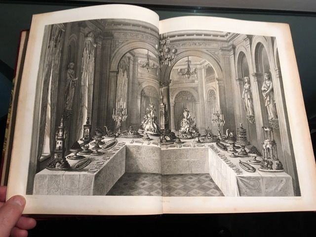 Un livre ouvert dévoile une gravure représentant une table de banquet ornée en forme de U, remplie de plats de service décoratifs et de statues.