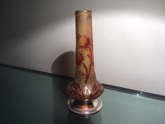 Un vase en verre ambré orné de motifs de chardon repose sur un socle en argent devant un mur uni.