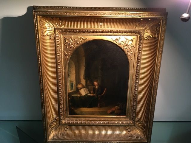 Tableau à l'huile représentant un érudit lisant à son bureau, dans une pièce sombre avec un globe terrestre, encadré d'une bordure dorée ornée.