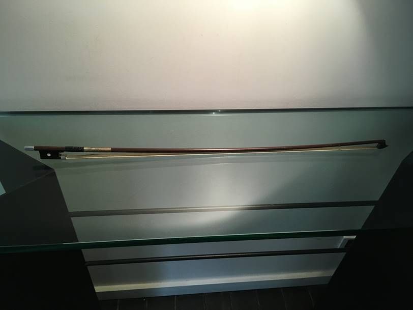 Un archet de violon en bois sombre repose horizontalement sur une étagère en verre, contre un mur clair et uni.