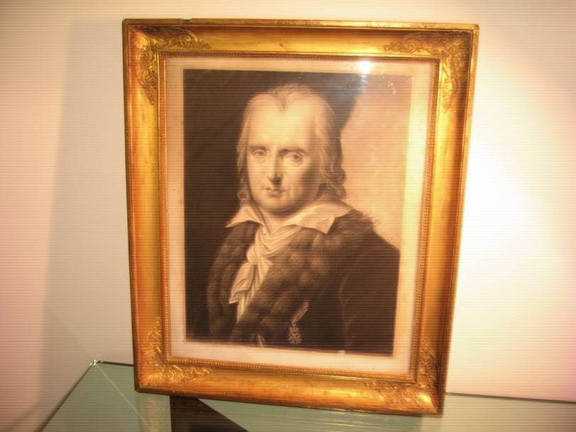Un portrait encadré, aux tons sépia, d'une personne portant un manteau à col de fourrure sur fond sombre, dans un cadre doré orné.