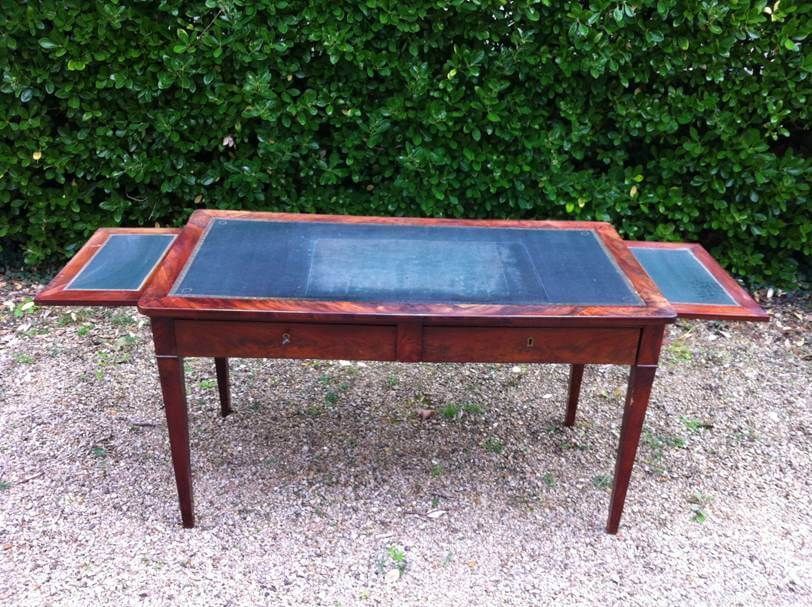 Bureau ancien en bois avec un plateau en cuir vert foncé et deux rallonges latérales, posé sur un chemin de gravier.