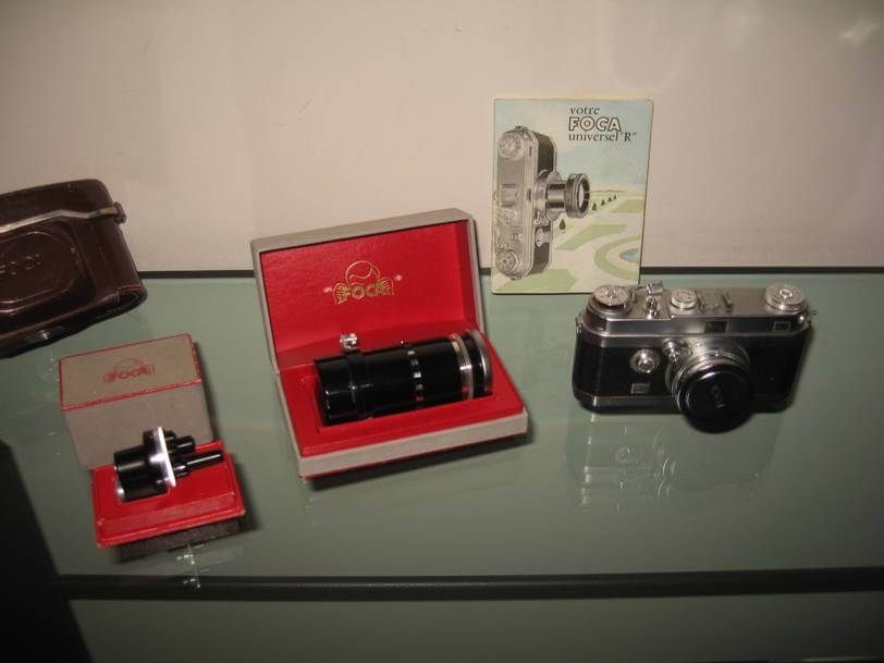 Appareil photo Foca vintage, accessoires et manuel présentés dans leurs boîtes d'origine sur une étagère en verre.