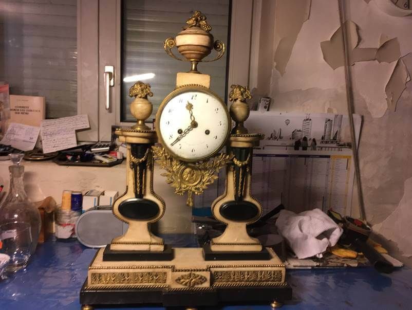 Une pendule de cheminée néoclassique ornée de détails crème et noirs, surmontée d'une petite urne décorative, posée sur un bureau encombré.