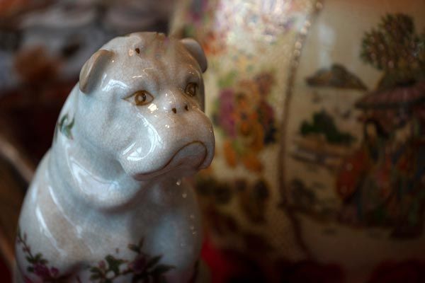 Gros plan d'une figurine de bouledogue en porcelaine blanche, placée à côté d'un vase flou à motifs floraux et paysagers.