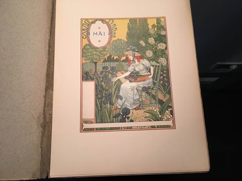 Une illustration ancienne intitulée MAI représente une personne en robe longue assise dans un jardin, s'occupant de fleurs de couleur sombre.