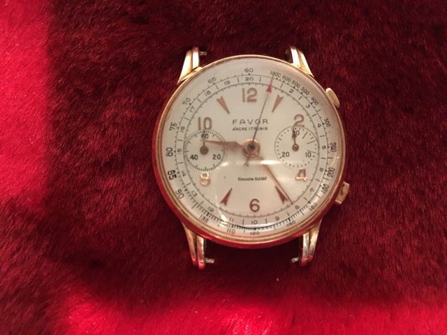 Une montre-bracelet chronographe Favor vintage couleur or, avec un cadran blanc et deux sous-cadrans, sur fond rouge foncé.