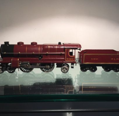 Une locomotive à vapeur miniature et son tender, peints en rouge foncé avec des garnitures dorées, reposent sur une surface en verre réfléchissant.