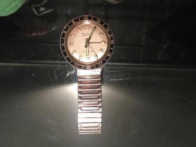 Une montre-bracelet analogique argentée, à cadran circulaire et lunette numérotée, posée sur une surface sombre et réfléchissante.