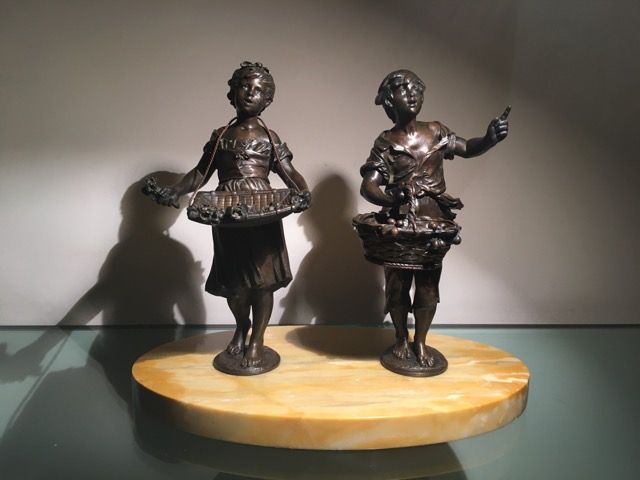 Deux statues en bronze d'enfants tenant des paniers se dressent sur un socle ovale en marbre jaune, devant un mur clair et uni.