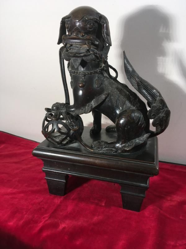 Une statue en bronze sombre représentant un lion gardien chinois tenant une boule, posée sur un piédestal devant un mur blanc et un tissu rouge.