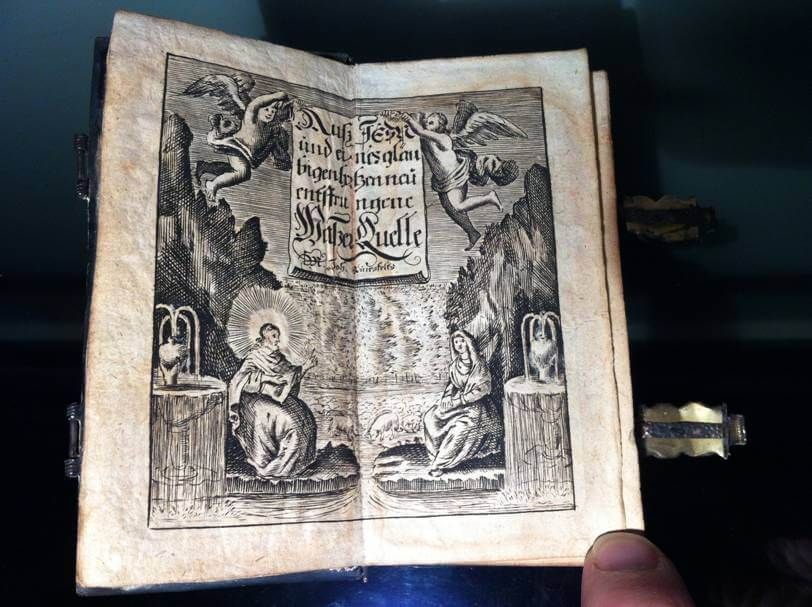 Un livre ancien ouvert, orné d'une illustration détaillée en noir et blanc représentant deux personnages, des anges, et un texte dans un rouleau central.