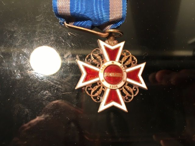 Médaille en forme de croix émaillée rouge et blanche, ornée d'une délicate filigrane d'or, suspendue à un ruban rayé bleu et gris.