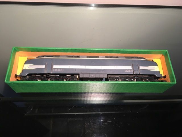 Une locomotive miniature aux couleurs bleues et grises, rangée dans une boîte de rangement rectangulaire verte avec un insert jaune.