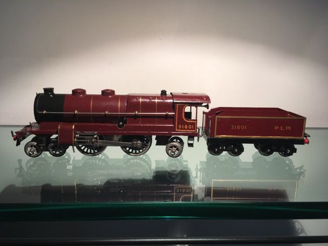 Une locomotive à vapeur miniature vintage de couleur bordeaux, avec son tender et la marque « PLM », est exposée sur une étagère en verre.