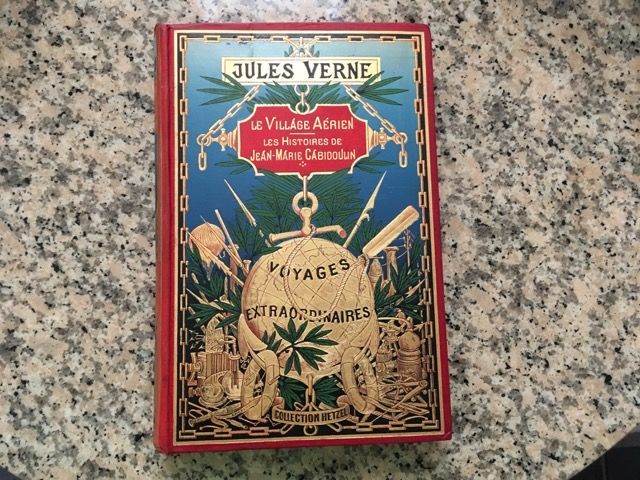 Une couverture de livre ornée, à la reliure rouge, de Jules Verne, intitulée Le Village Aérien, représentant un globe doré et des motifs maritimes.
