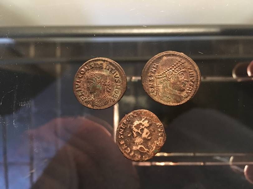 Trois pièces de monnaie romaines en bronze, ornées de portraits d'empereurs, disposées en triangle sur une surface sombre et réfléchissante.