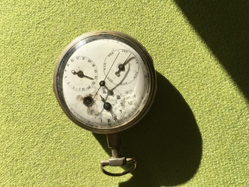 Une montre de poche ancienne, au cadran fissuré et à double cadran, repose sur une surface verte texturée sous un soleil éclatant.