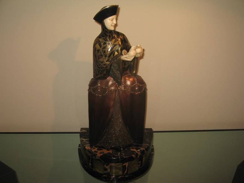 Sculpture représentant une figure vêtue d'une tunique sombre à motifs et d'une large jupe, se tenant sur un socle arrondi en marbre sombre.