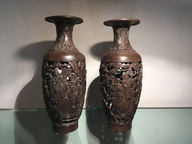 Deux vases en métal sombre, richement sculptés et ajourés, sont disposés côte à côte sur une surface en verre réfléchissante.