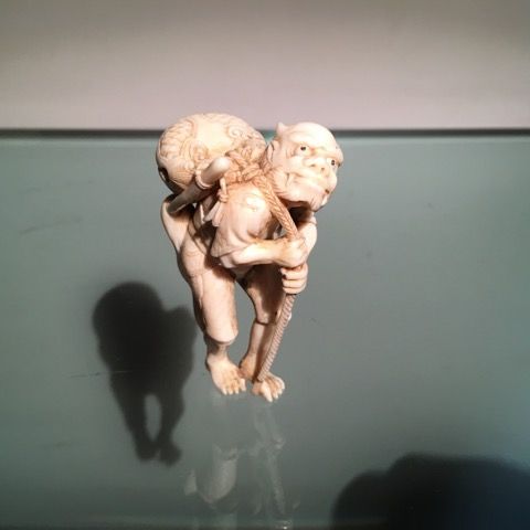 Un netsuke sculpté en ivoire représentant une figure courbée portant un grand sac rond sur le dos tout en tenant une corde.