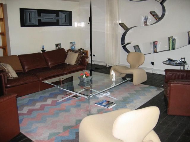 Salon avec un canapé en cuir marron, deux fauteuils modernes beiges, une table basse en verre et une étagère murale ondulée.