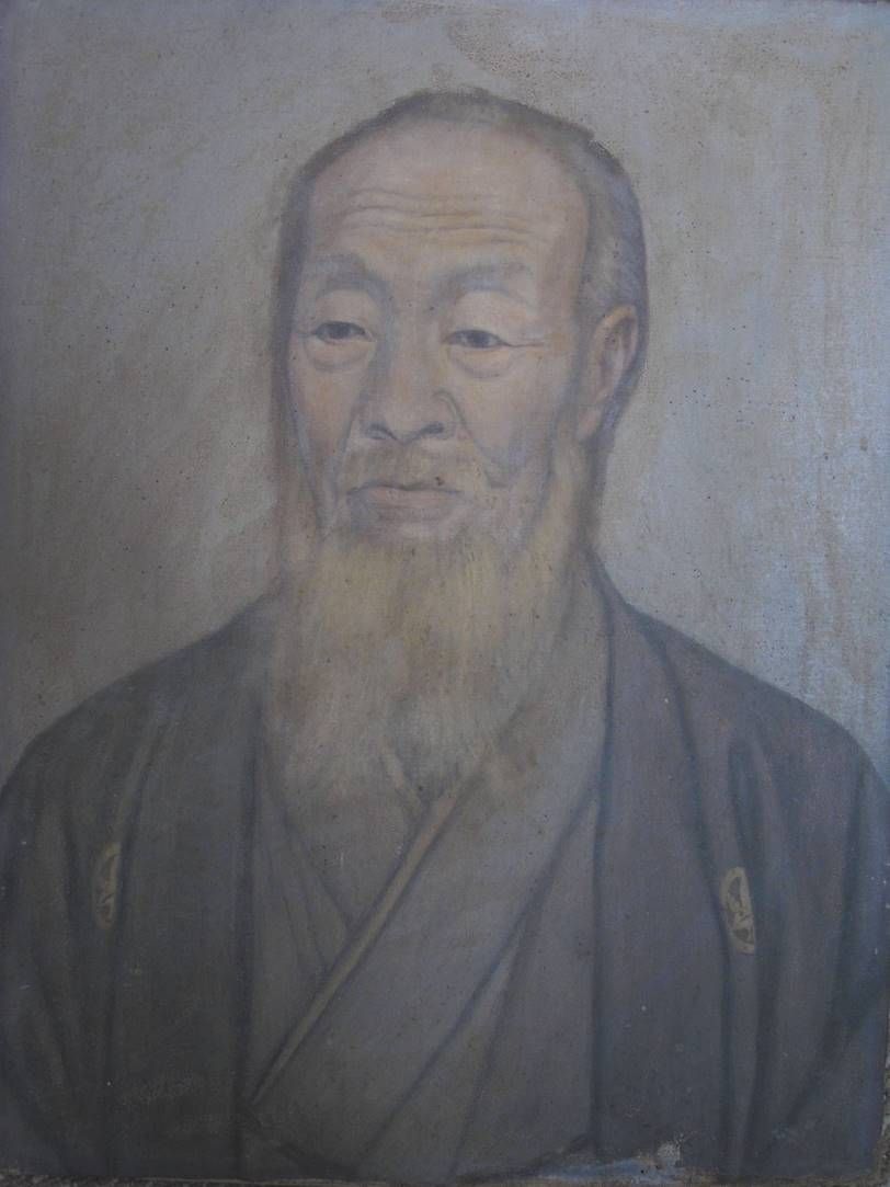Portrait d'une personne à la longue barbe, vêtue d'un vêtement traditionnel sombre, sur un fond uni aux tons clairs.