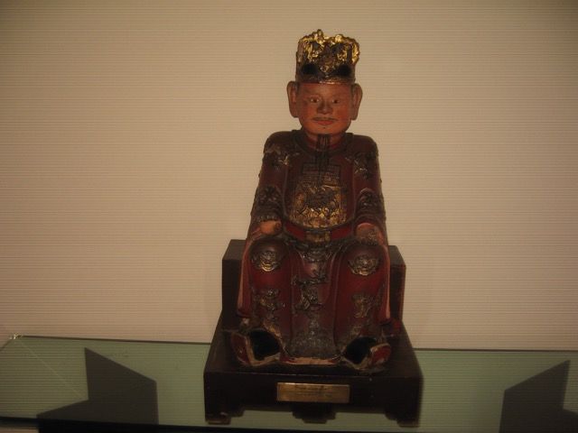 Une figure assise, rouge et or, portant une couronne traditionnelle, placée sur un socle en bois sombre sur fond uni.