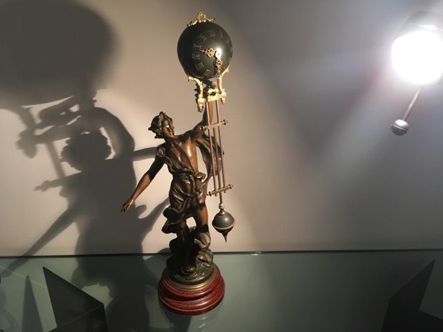 Une sculpture en bronze représente une figure tenant une horloge ronde suspendue par une structure ornée au-dessus d'une base circulaire, projetant une grande ombre.