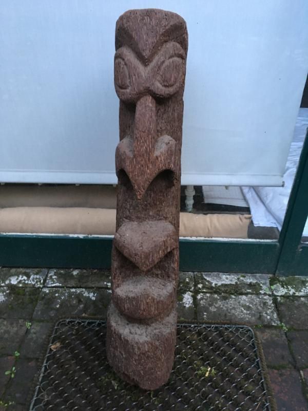 Une statue tiki en bois sculpté, au visage sévère et stylisé, dressée sur une grille métallique.