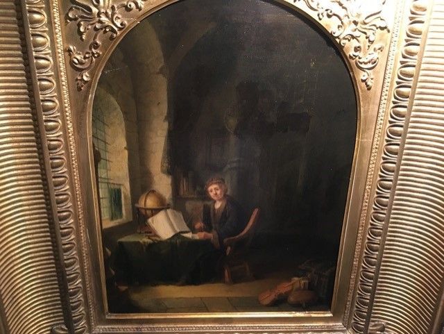 Une personne est assise à un bureau dans une pièce sombre et voûtée, éclairée par une fenêtre, en train de lire un livre à côté d'un globe terrestre.