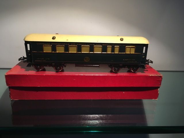 Un wagon de train miniature ancien, avec une caisse verte et un toit crème, posé sur une boîte rouge sur une surface en verre.