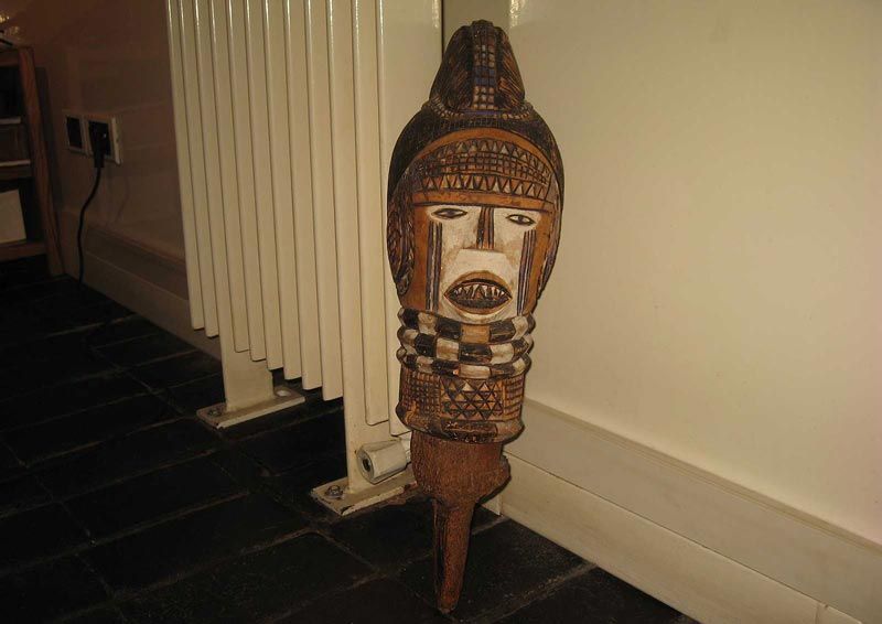 Un masque en bois finement sculpté, orné de motifs géométriques et présentant une bouche ouverte, est appuyé contre un radiateur blanc.