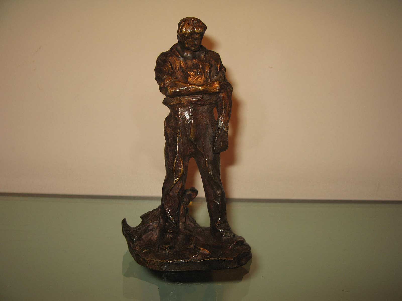 Une petite sculpture de couleur bronze représentant un personnage debout, les bras croisés, sur un fond clair uni.
