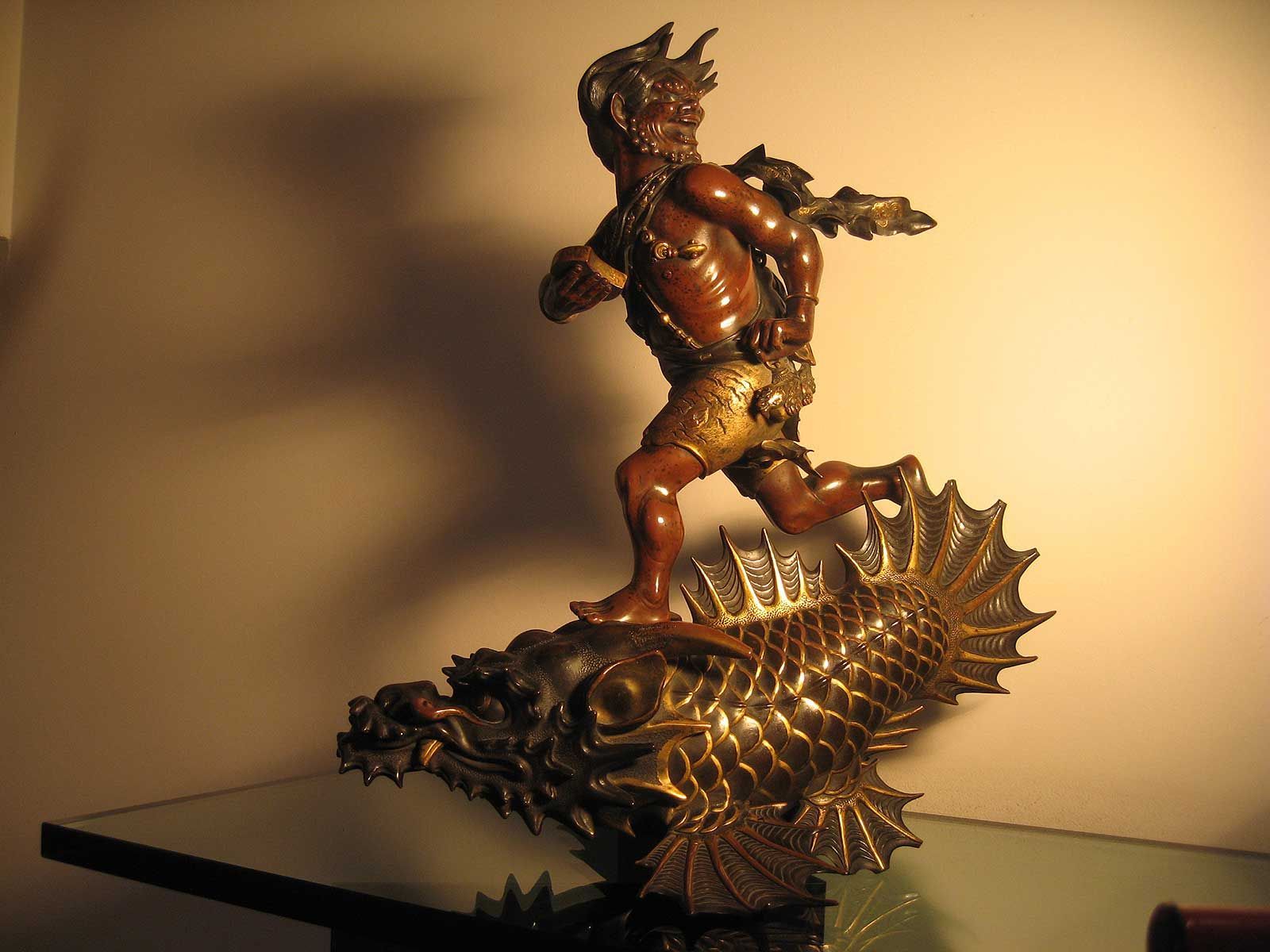 Une sculpture en bronze représente une figure mythologique courant sur un poisson stylisé à écailles, devant un mur clair et uni.