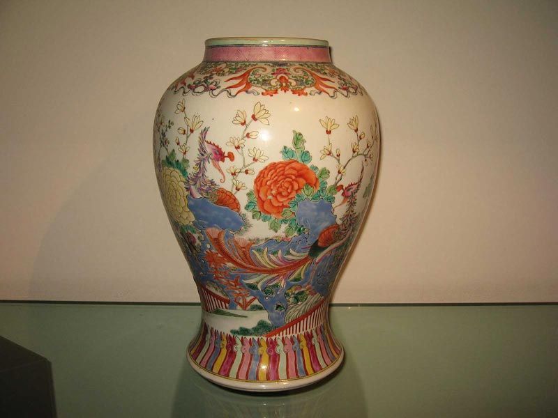 Un vase en céramique à col et base fuselés, orné de motifs floraux colorés et de créatures stylisées sur fond blanc.