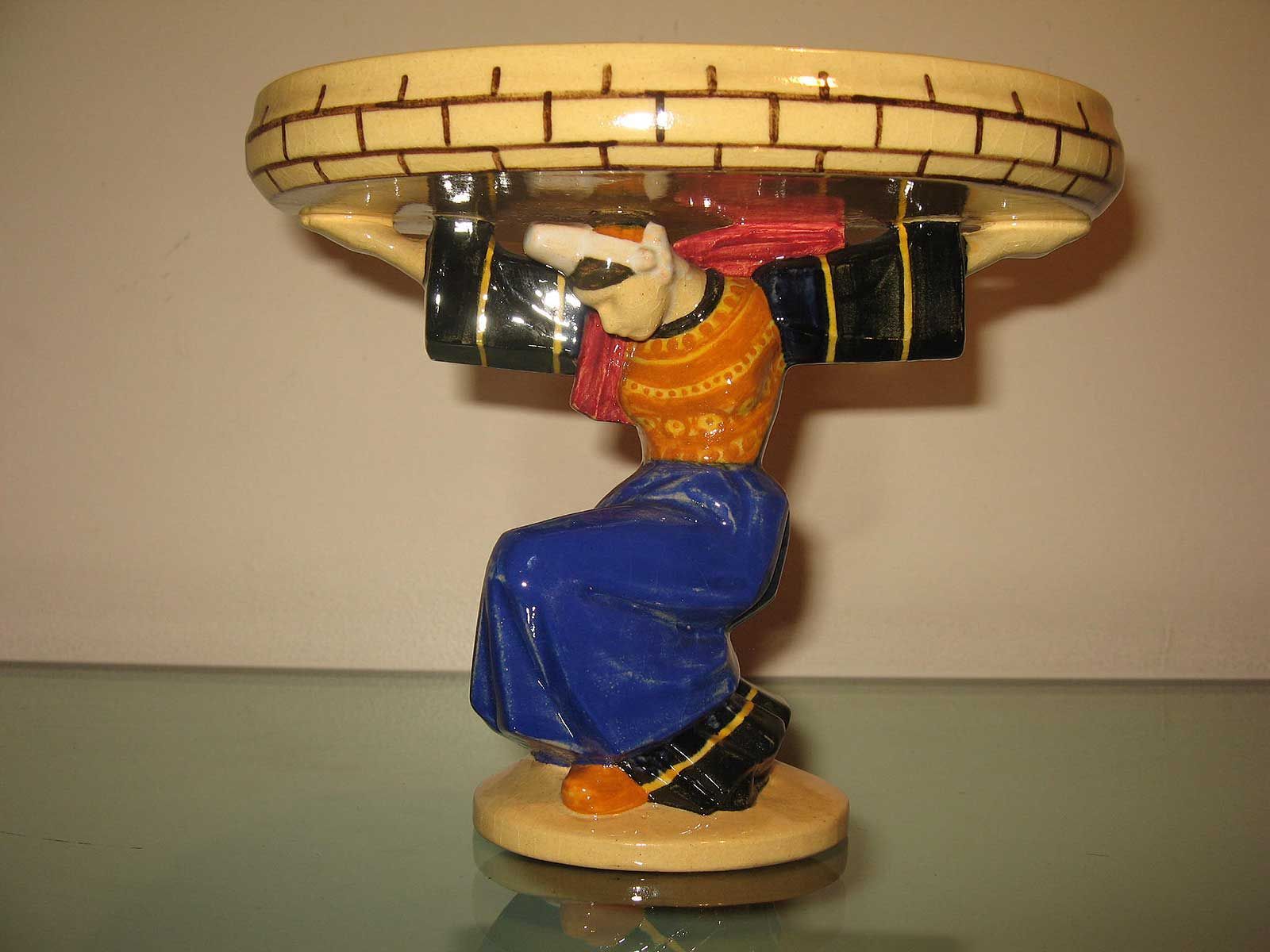 Un bol sur piédestal en céramique peint à la main, représentant un personnage accroupi soutenant sur sa tête un rebord à motif de briques.