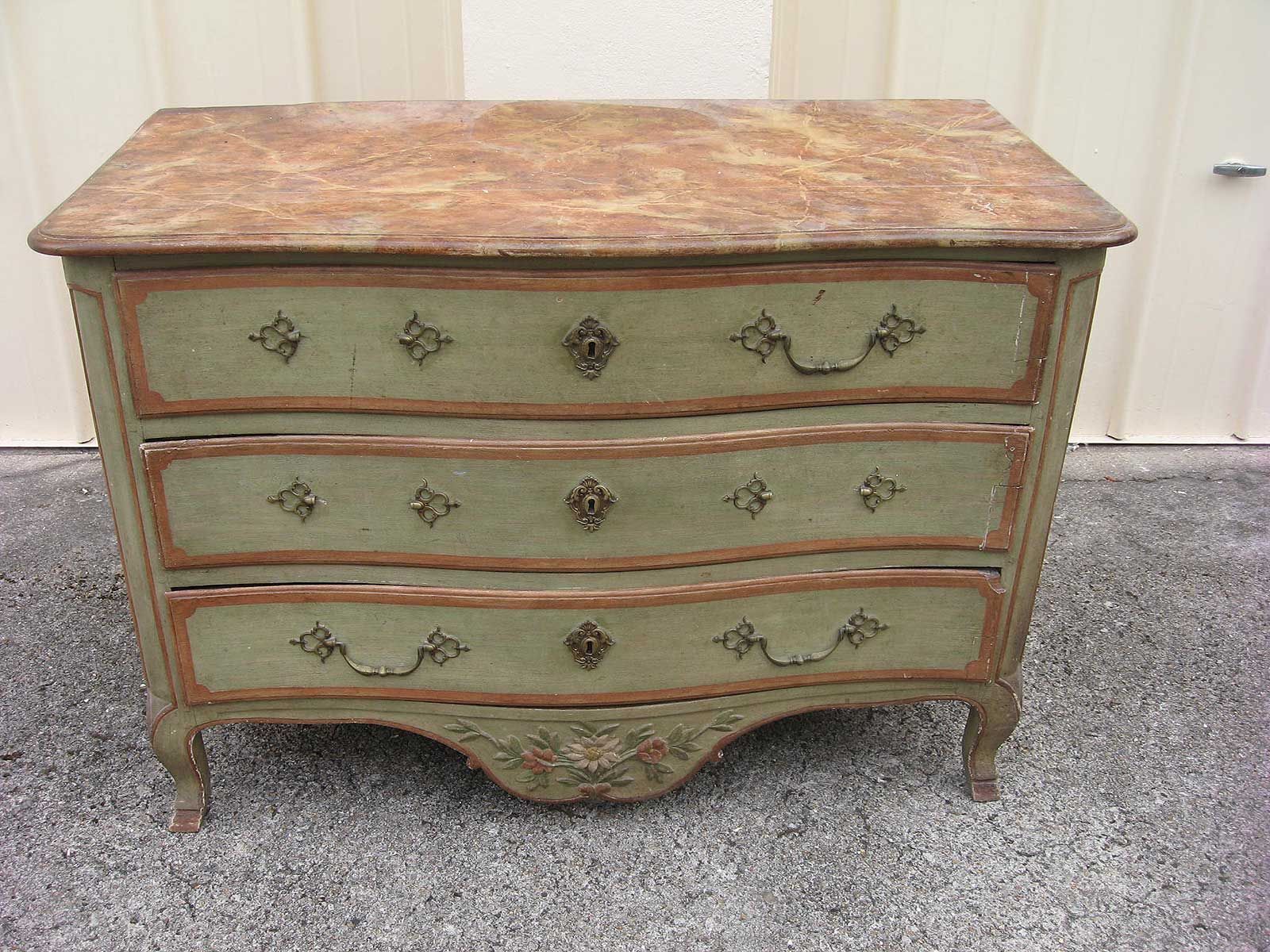 Une commode en bois vert sauge vieilli, avec trois tiroirs, des ferrures en laiton et un plateau en marbre brun moucheté.