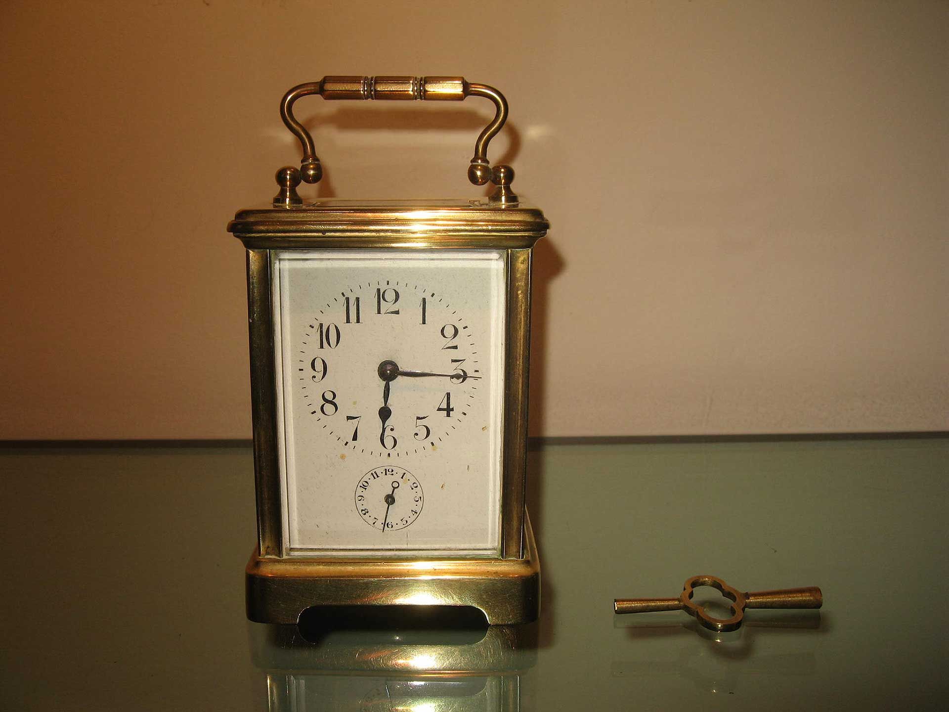 Une pendule de voyage en laiton, à cadran blanc et petite clé, posée sur une surface en verre sur fond clair.