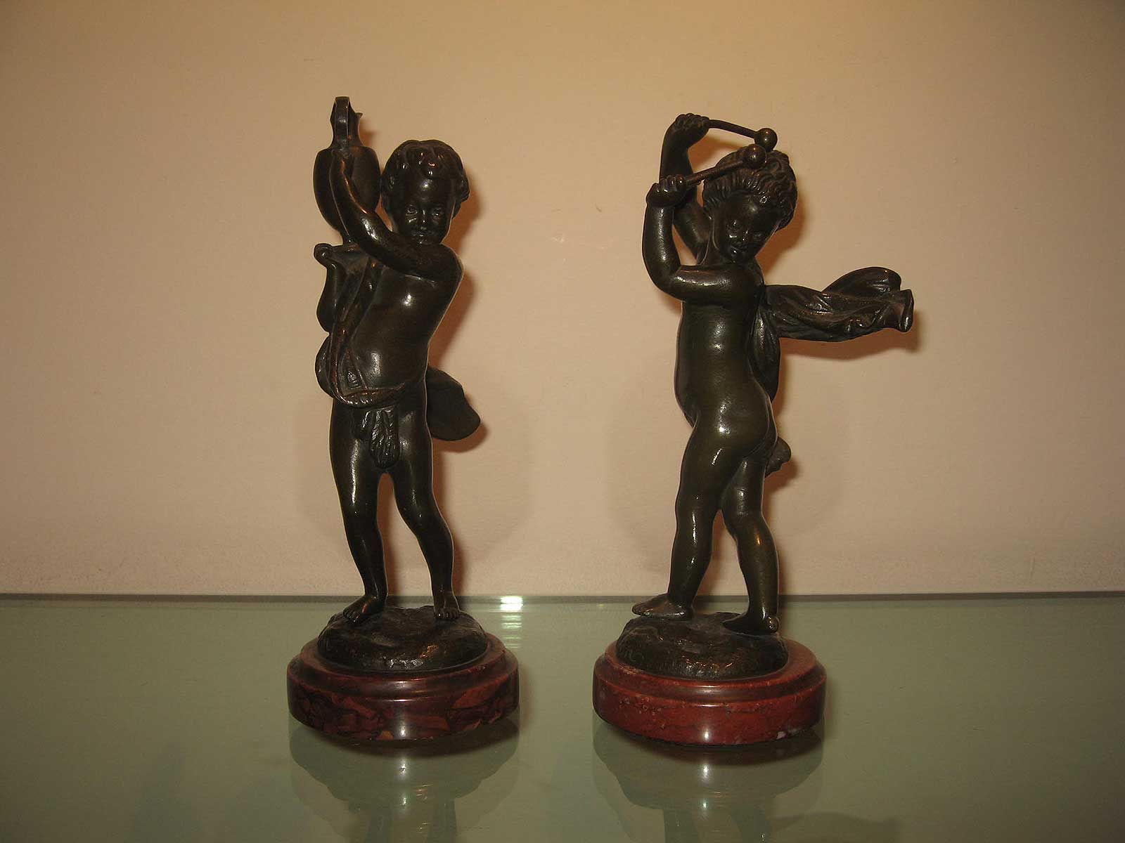 Deux statues de chérubins en bronze sombre sur des socles circulaires en bois, l'une tenant un oiseau et l'autre un petit marteau.