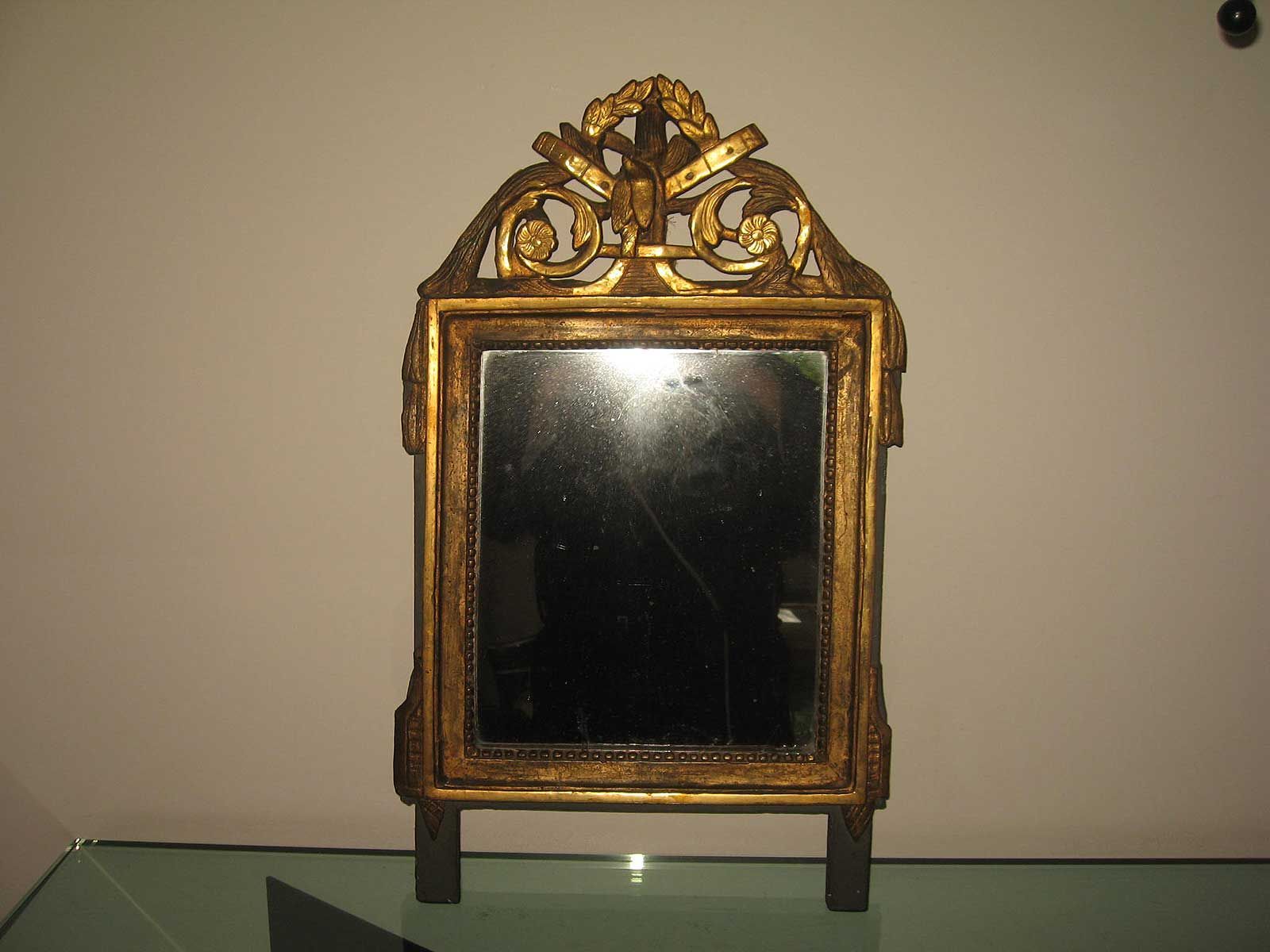 Un miroir rectangulaire orné, à cadre doré et décoré de volutes sculptées en partie supérieure, posé sur une surface en verre.