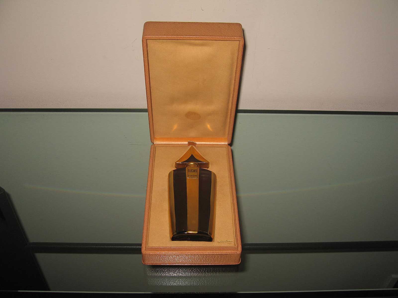 Un briquet doré en forme de flèche, posé dans un coffret de présentation doublé de tissu beige sur une surface réfléchissante.