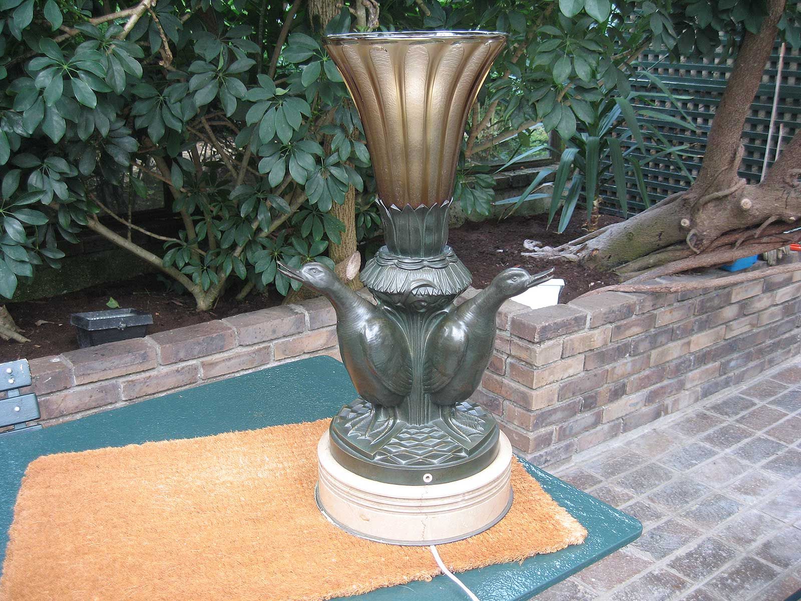 Une fontaine extérieure couleur bronze ornée de trois figurines de canards autour d'un bassin cannelé en forme de trompette, sur une terrasse en briques.