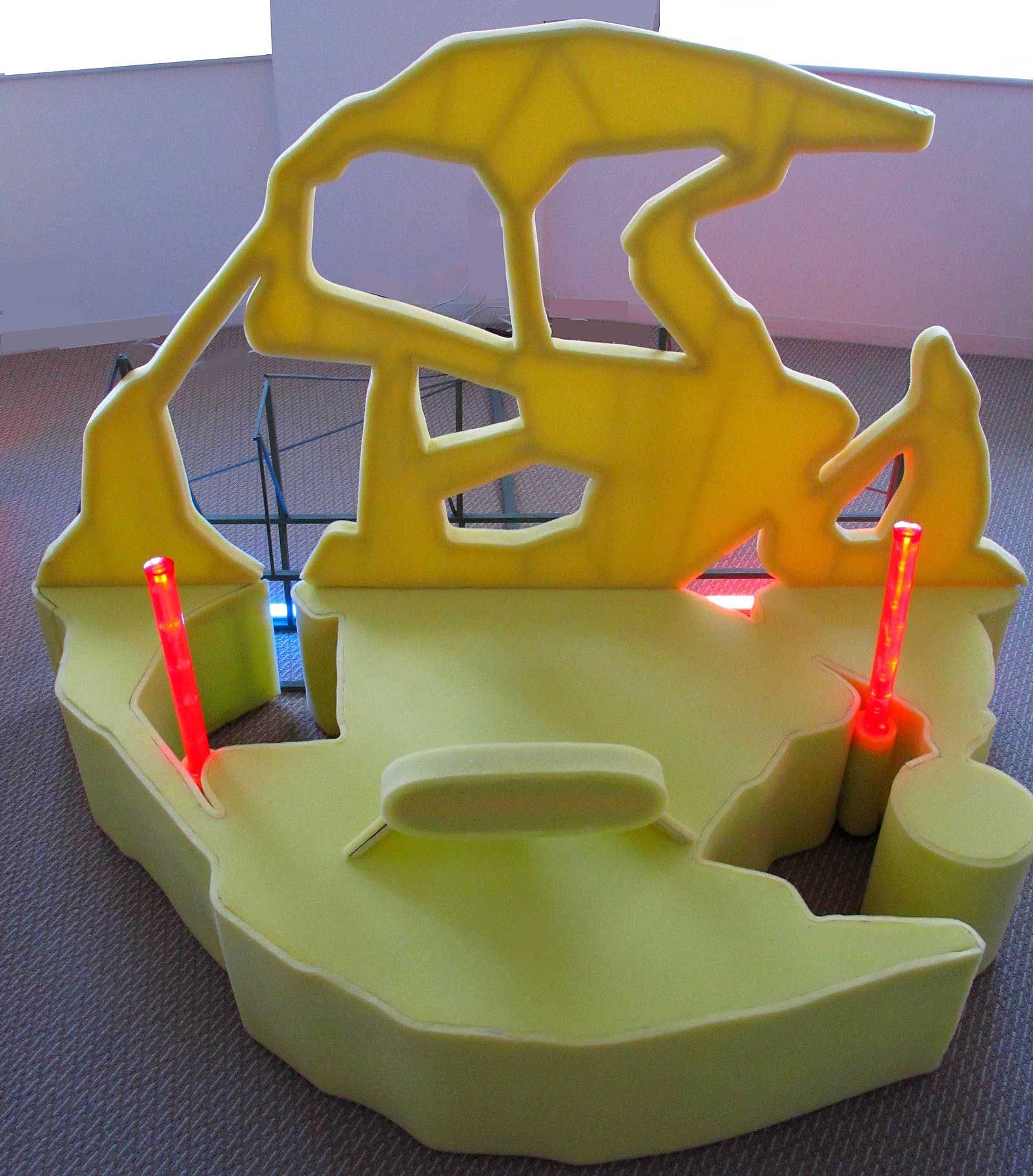 Un ensemble de sièges modulaires en mousse jaune vif, orné de découpes abstraites et de deux piliers lumineux rouges verticaux, sur un sol recouvert de moquette.
