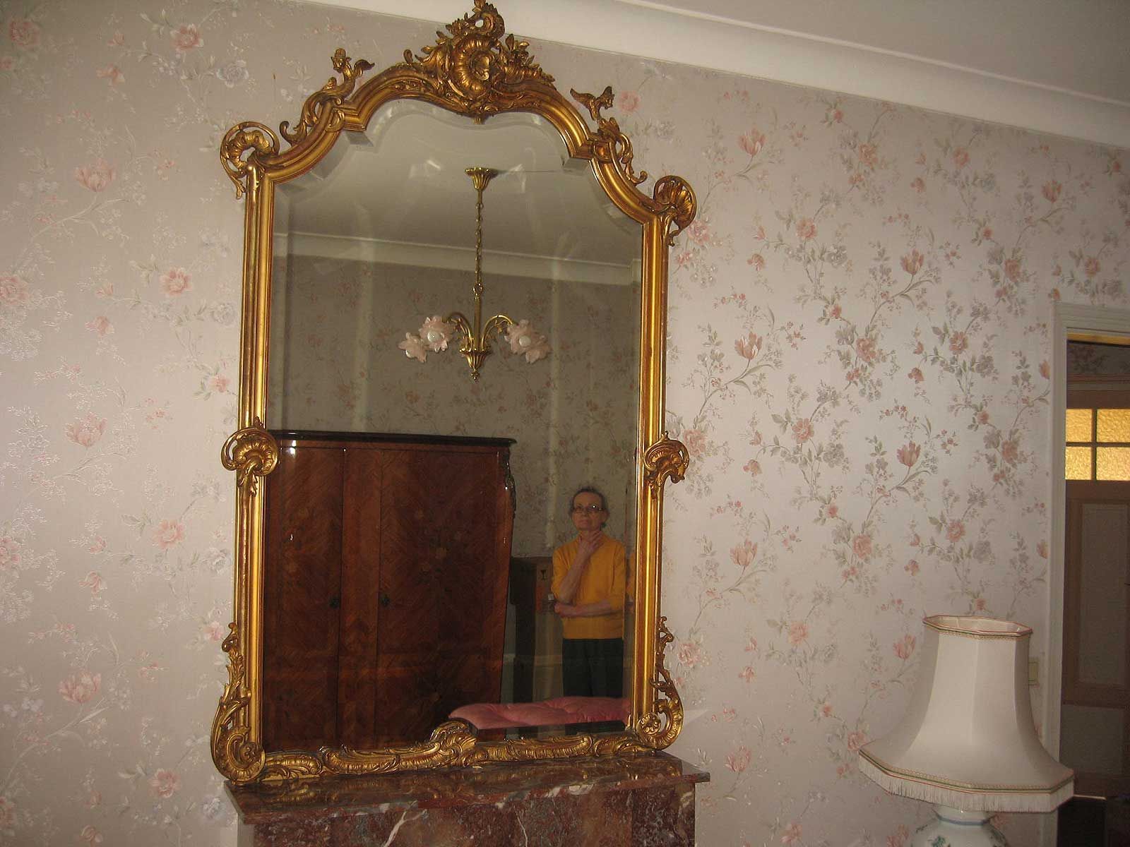 Un miroir orné d'un cadre doré reflète l'intérieur d'une pièce et une personne se tenant devant lui, sur un fond de papier peint à motifs floraux.
