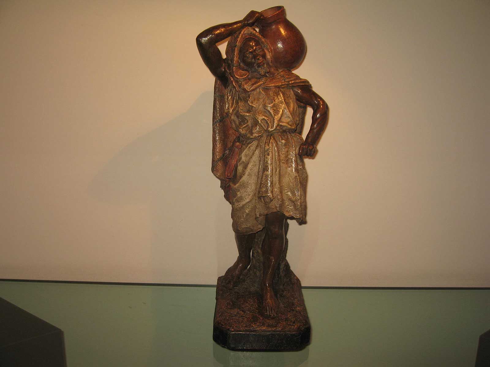 Une sculpture de couleur bronze représentant une personne debout, vêtue de vêtements drapés et portant en équilibre une jarre d'eau ronde sur son épaule.
