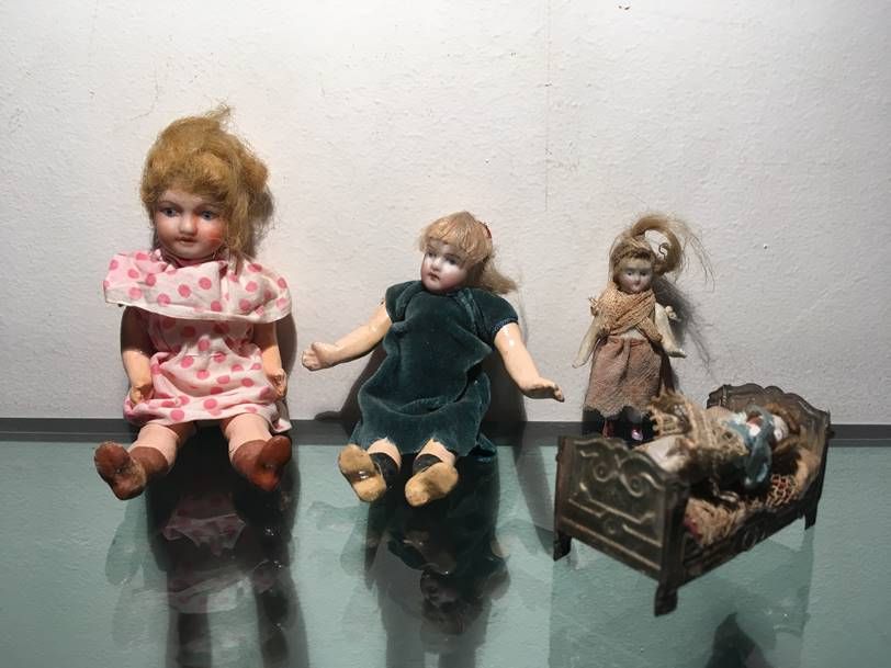 Trois poupées anciennes sont exposées sur une surface en verre, dont une assise dans un petit cadre de lit en métal.