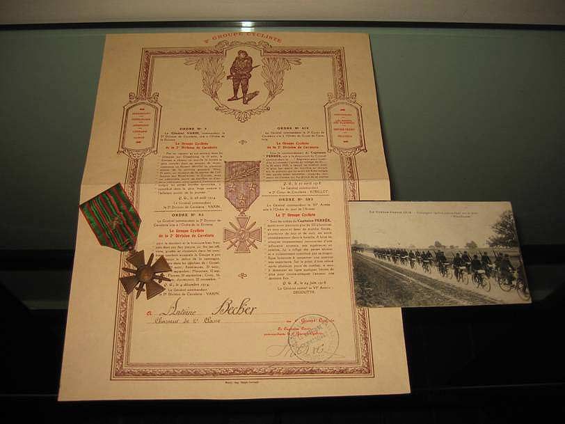 Un document commémoratif de décoration militaire datant de la Première Guerre mondiale, une médaille en forme de croix à suspendre et une carte postale ancienne montrant des soldats dans un paysage.