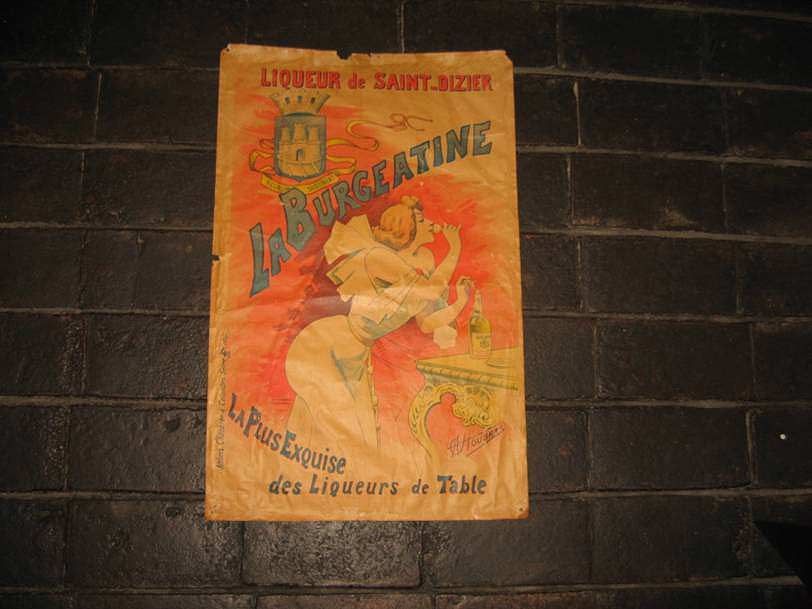 Affiche vintage pour la liqueur La Burgeatine sur un mur de briques sombres, représentant une femme à une table ornée d'un logo de château.